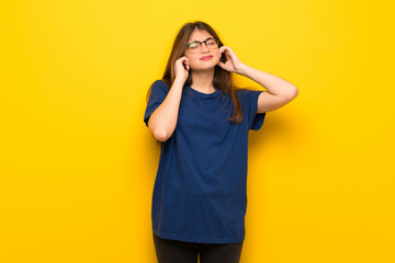 Young woman with glasses over yellow wall covering ears with hands. Frustrated expression