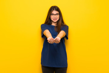 Young woman with glasses over yellow wall holding copyspace imaginary on the palm to insert an ad