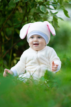 Infant girl in rabbit suit seating on grass in park and smiling.