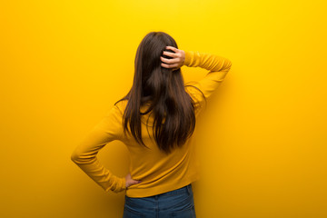Teenager girl on vibrant yellow background on back position looking back while scratching head
