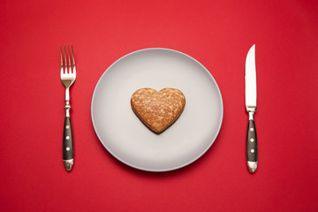 Heartbreaker concept image: heart shaped cookie on plate with fork and knife over red background