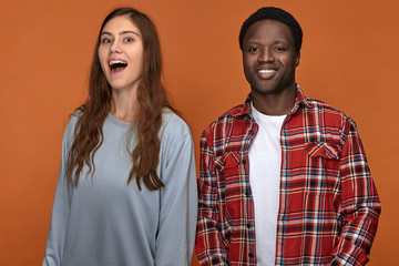 Isolated shot of attractive dark skinned guy in stylish clothes having fun with his charming female friend of Caucasian appearance, laughing, looking at camera, playing fools. Interracial couple