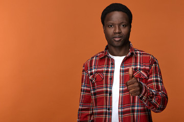 Portrait of stylish trendy looking young Afro American man in his twenties posing against blank studio background with thumbs up gesture, expressing approval, satisfaction and positive attitude