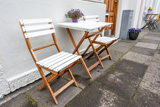 White Empty Wooden Table Outside Restaurant Cafe With Two Chairs On Sidewalk Street And Purple Flowers In Flowerpot Potted Plant Setting With Nobody In Iceland