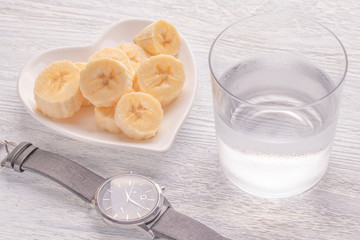 Sliced and whole bananas. On a white plate. On the old wooden table. Misted glass with clear water. Wrist Watch
