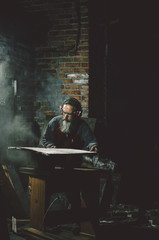 Old bearded carpenter woodworker  cutting wood plate with circular saw, process of working, rustic old loft dark style