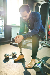 Naklejka premium Sad amputee young bearded man sitting on sort bench and leaning on knees in health club, small dumbbells on floor