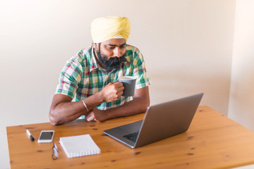 Indian with turban working with his laptop and  holding a cup of coffee