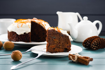 Delicious chocolate fruit cake ornated with cream icing and orange peel served with tea, nuts, cones and cinnamon sticks on blue background