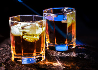 Two glasses of the whiskey on the natural stone plate with blue laser ray and shadows from the glasses