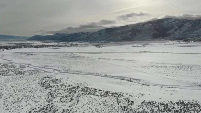 Aerial-Flying over snowy field dotted with sagebrush toward setting sun and shadowy mountains over snow-filled creek bed