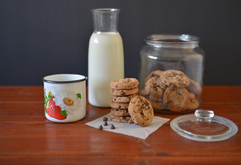 chocolate chip  cookies in glass jar with glass bottle of milk and white enamel mug on wooden rustic background