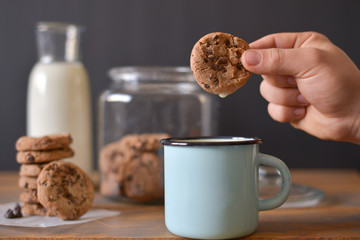 chocolate chip  cookies in glass jar with glass bottle of milk and turquoise enamel mug on wooden rustic background with men hand holding one cookie 