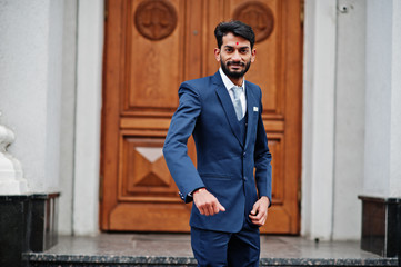 Stylish beard indian man with bindi on forehead, wear on blue suit posed outdoor against door of building.