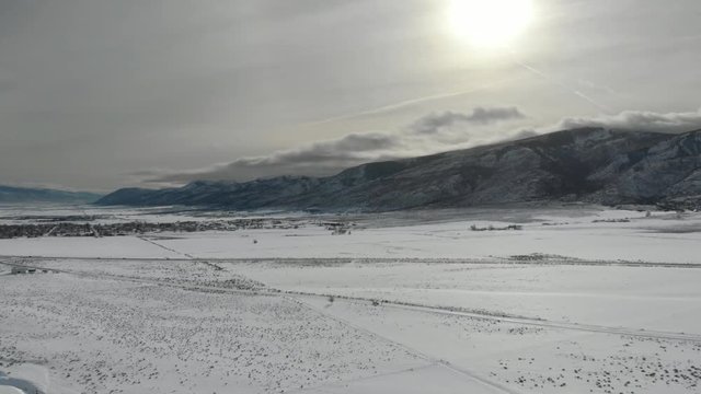 Aerial-Flying high over snowy field toward setting sun and shadowy mountains under hazy cloudy sky