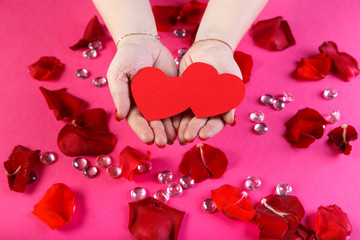 Red double paper heart in female hands on a pink background surrounded by rose petals.