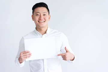 Smiling Asian man holding blank sheet of paper. Young guy pointing at paper sheet and looking at camera. Promotion concept. Isolated front view on white background.