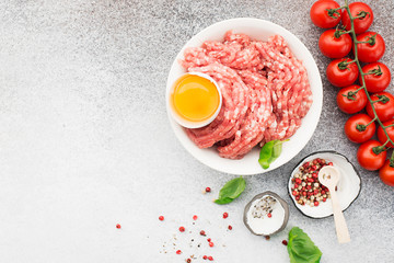 Fresh minced pork with egg, salt, pink pepper, cherry tomatoes on a light background with basil leaves. Top view.
