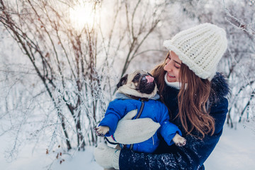 Pug dog walking on snow with his master. Puppy wearing winter coat. Woman hugging his pet in winter forest