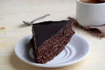 The piece of chocolate cake  is on a white plate. The cup with coffee is near cake.