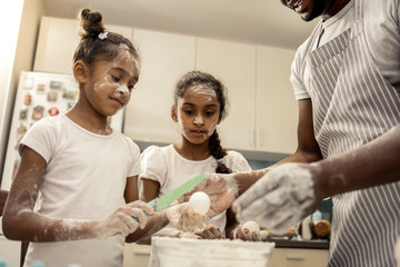 Dark-haired daughters with faces in flour helping father in kitchen
