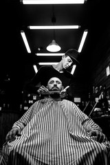 Fototapeta na wymiar Black and white. The stylish barbershop. The fashion barber tidies up beard of brutal man sitting in the armchair