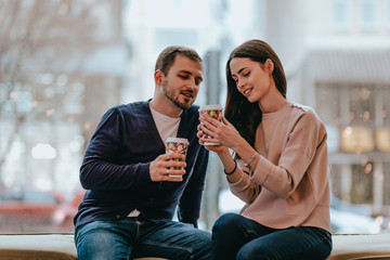 Lovers guy and girl dressed in sweaters and jeans are sitting close to each other on the windowsill in a cafe and drinking from cups