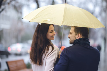 Happy romantic couple, guy and his girlfriend dressed in casual clothes walk under the umbrella and look at each other on the street in the rain.