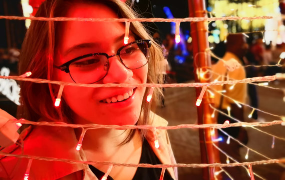 Fototapeta Hispanic young woman with white skin and beautiful smile at a fair of lights at Christmas. Abstract red color on his face. Detail of her hair and big black frame glasses.