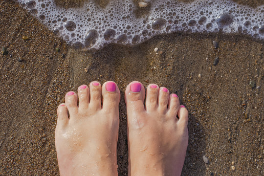 Top view photography of beautiful female barefeet legs with professional pink painted toenails. Pink color pedicure. White tanned woman standing and relaxing on sandy beach at seashore on summer day.