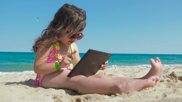 Funny kid using tablet on the beach. A beautiful little girl in a swimsuit in sunglasses and a tablet sits on the sand on a beach against the backdrop of the sea.