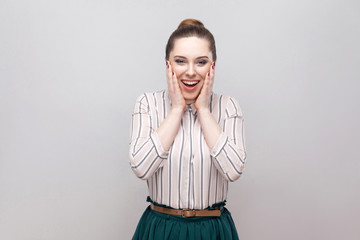 Fototapeta premium Portrait of happy surprised beautiful young woman in striped shirt, collected ban hairstyle, standing, amazed and looking at camera with toothy smile. indoor studio shot, isolated on grey background.