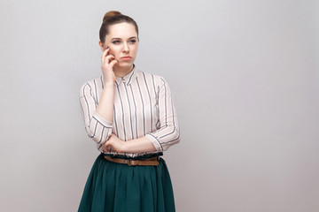 Confused beautiful young woman in striped shirt and green skirt with makeup and collected ban hairstyle, standing thoughtful and looking away. indoor studio shot, isolated on grey background.