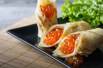 horizontal photo of the black plate with pancakes filled by red caviar and salad leaves 