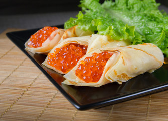horizontal photo of the black plate with pancakes filled by red caviar and salad leaves 