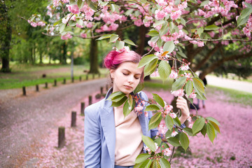 Fototapeta premium Beautiful girl with pink hair in a fashionable blue pantsuit standing next to colorful flowers.