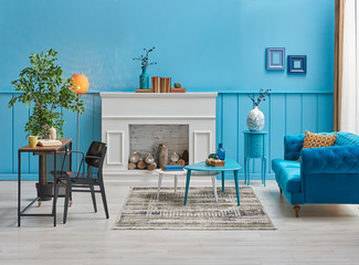 Decorative blue wall, white fireplace and blue sofa. Wooden desk and black chair in the room with lamp and vase of plant.