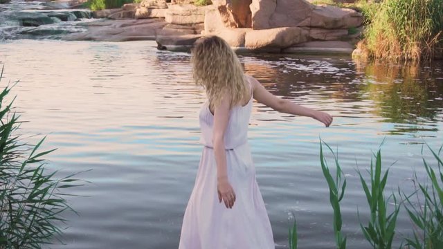 positive blonde girl barefoot dancing on a stone by the river like a fabulous mermaid, her pink long gentle hipster dress flies and flutters, sun glare on the water on a bright warm day