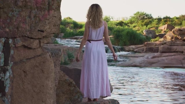 slender tall girl with curly bloody hair stands on a big namke near a huge rock and raises her arms slightly, dressed in a summer pink dress, as if a mermaid preparing to jump into the water, no face