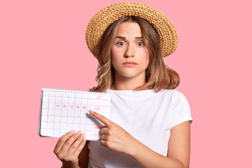 Horizontal shot of pleasant looking woman with appealing look, holds periods calendar for checking menstruation days, points with fore finger, isolated over pink background. Gynecological concept