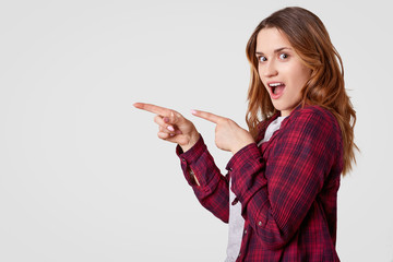 Horizontal shot of cheerful young woman with long hair, points with both fore fingers at empty space, wears casual checkered shirt, shows direction to somewhere, isolated over white background.