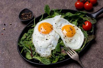 Fried eggs sunny side up on pan with herbs, cherry tomatoes and pepper, with fork an brown background. Concept of healthy breakfast