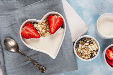 Yogurt with muesli and strawberries in a heart-shaped bowl on a pastel blue background.