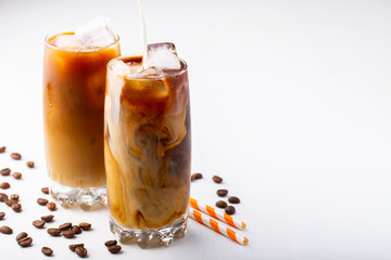 Ice coffee in a tall glass with cream poured over and coffee beans. Cold summer drink on white background with copy space