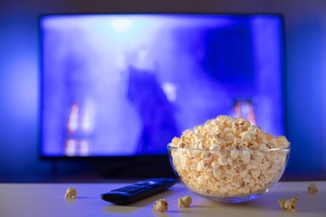 A glass bowl of popcorn and remote control in the background the TV works. Evening cozy watching a movie or TV series at home