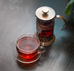 hot black tea with steam over it in a Cup and a glass copper teapot next to it on a wooden table