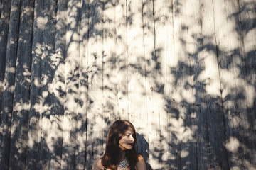 Stylish hipster girl posing in sunny street on background of wooden wall. Boho girl in cool outfit standing in sunlight and shadow. Space for text. Summer vacation and travel. Creative photo