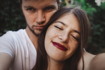 Naklejka premium Stylish hipster couple making selfie and embracing. Happy family couple in love making self portrait and smiling in evening summer city. Vacation and travel together