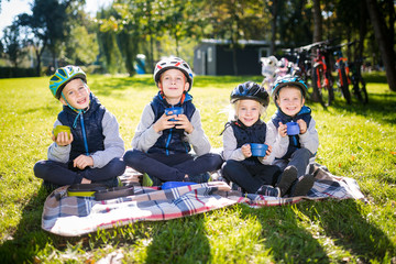 Theme active family holidays nature. group people small little children three brothers and sister sit onblanket near bicycles in park green grass lawn rest and drink drink from cups and thermos tea