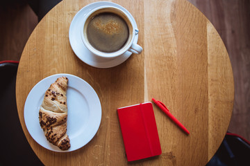 A red note and red pen is on the table in a cafe, a cup of coffee and a croissant